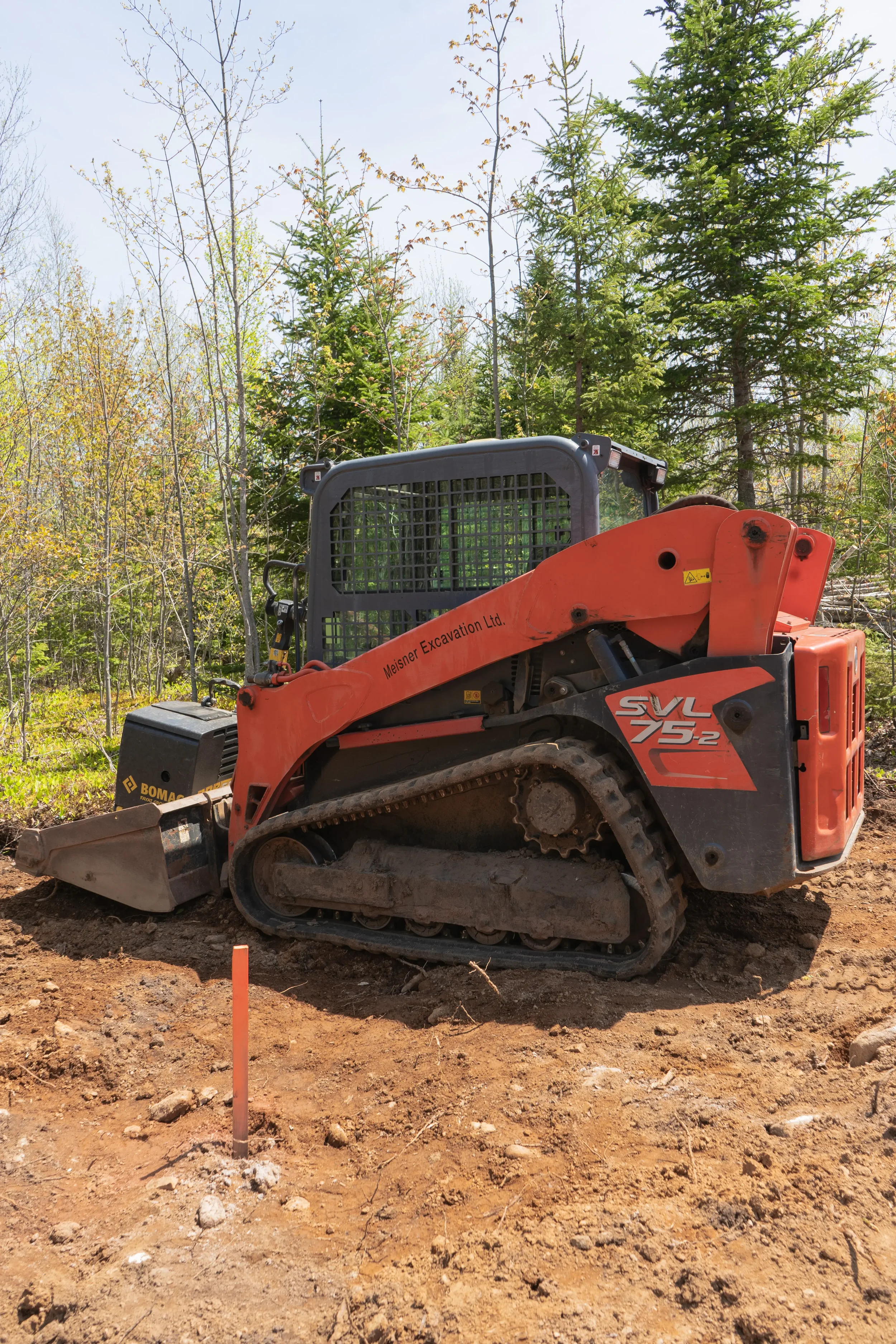 A small orange and black tracked excavator on dirt, surrounded by green trees and forest.