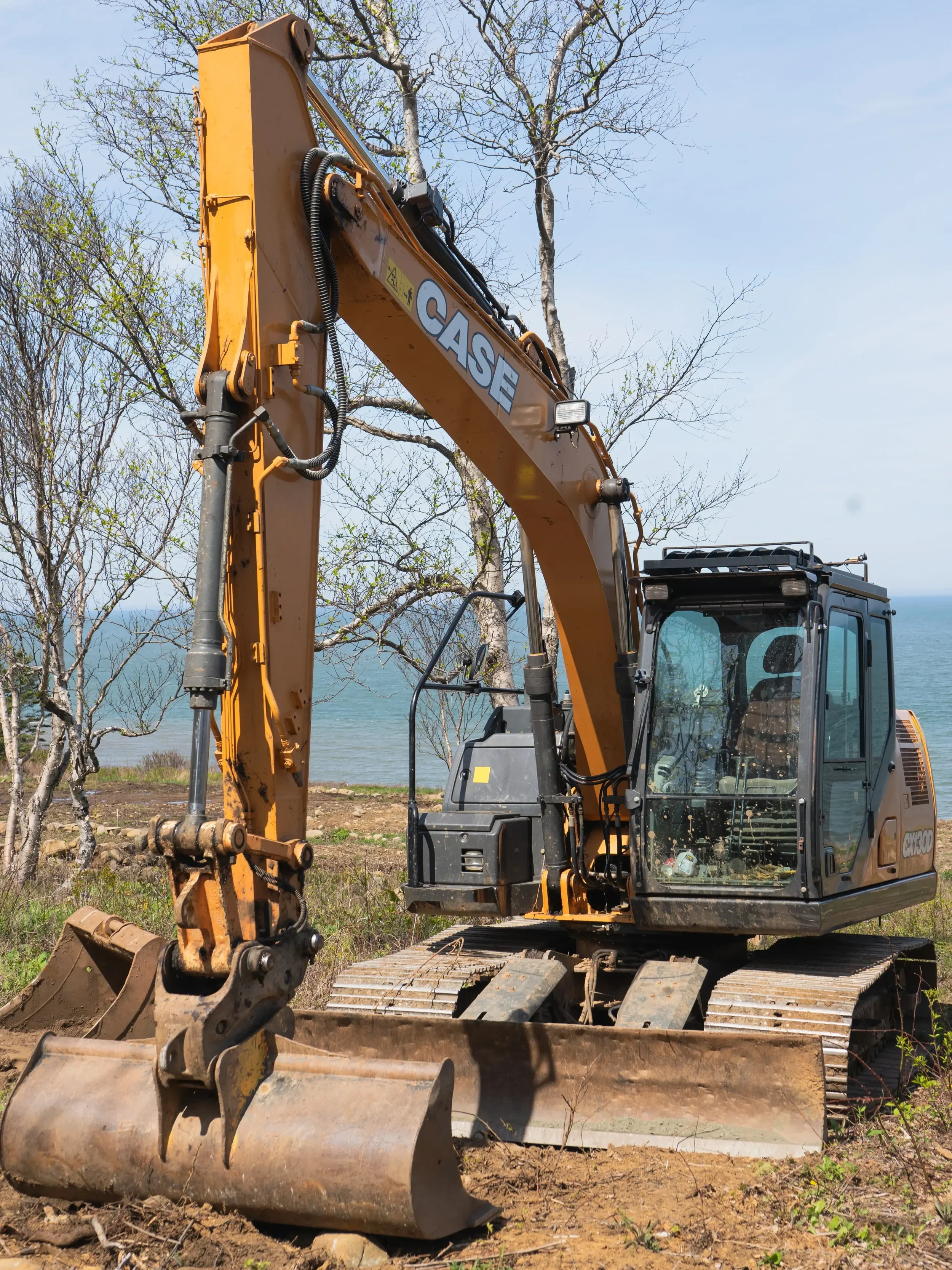 A yellow excavator on rocky terrain near a body of water with trees and a blue sky in the background.