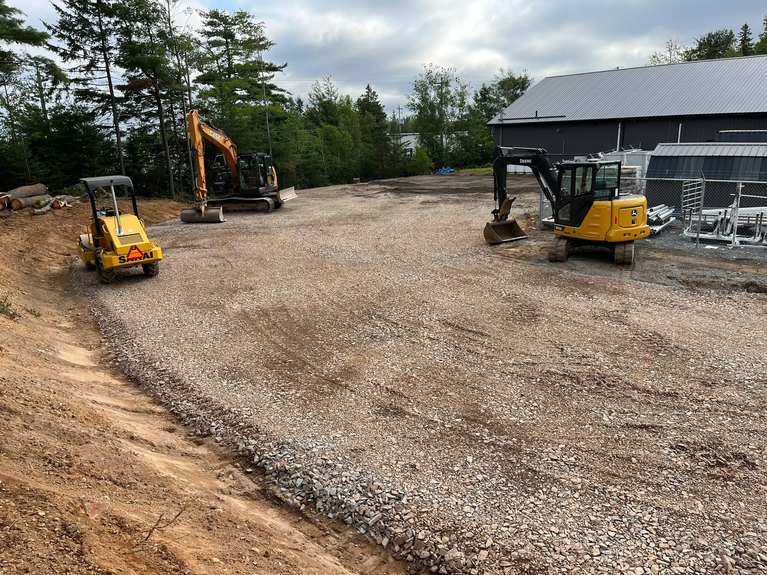 Construction site with a small roller, an excavator, and a mini excavator on gravel ground, with a building and trees in the background.