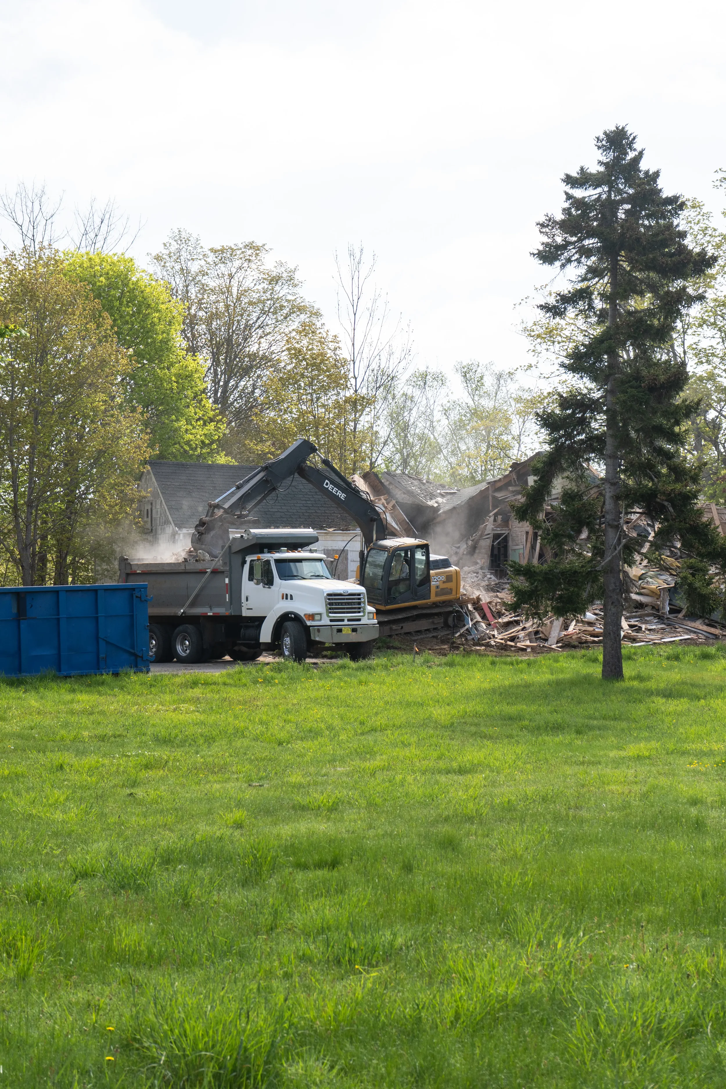 Construction workers demolishing a house using an excavator and a dump truck in a suburban area with trees and grassy yard.