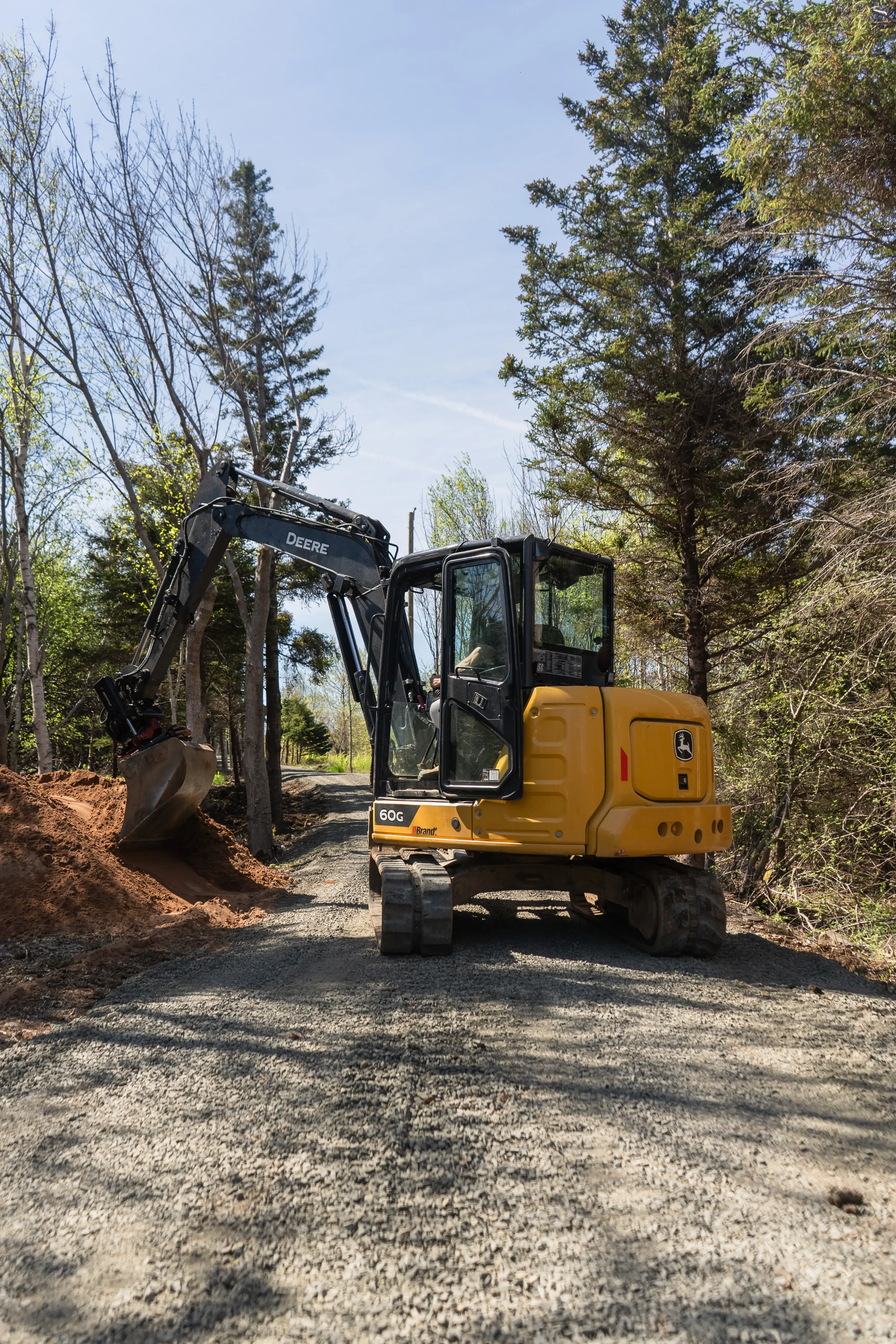 A yellow John Deere compact excavator working on a dirt path in a wooded area.