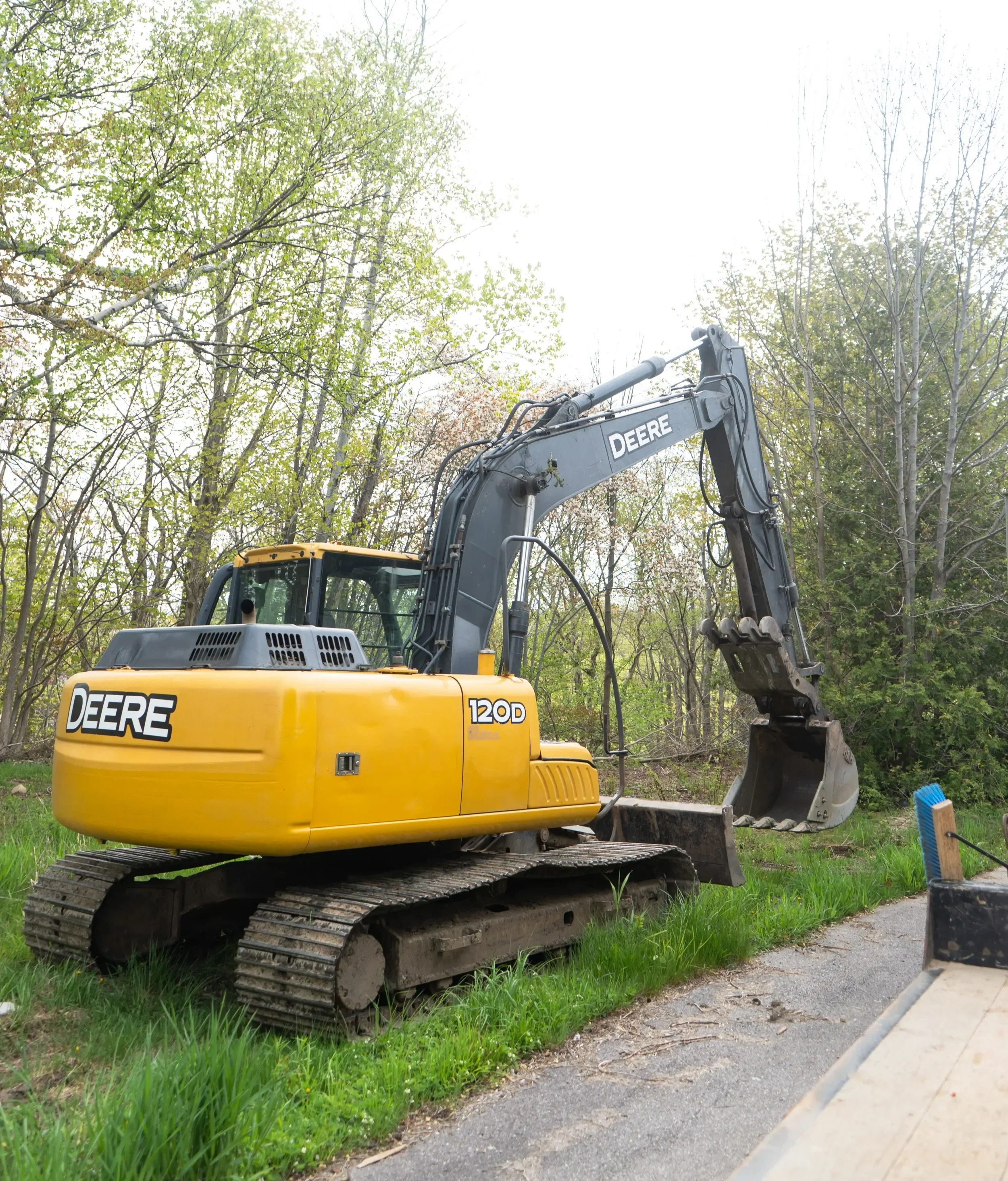 A yellow Deere 120D excavator parked on a patch of grass near a paved road with a wooded background.