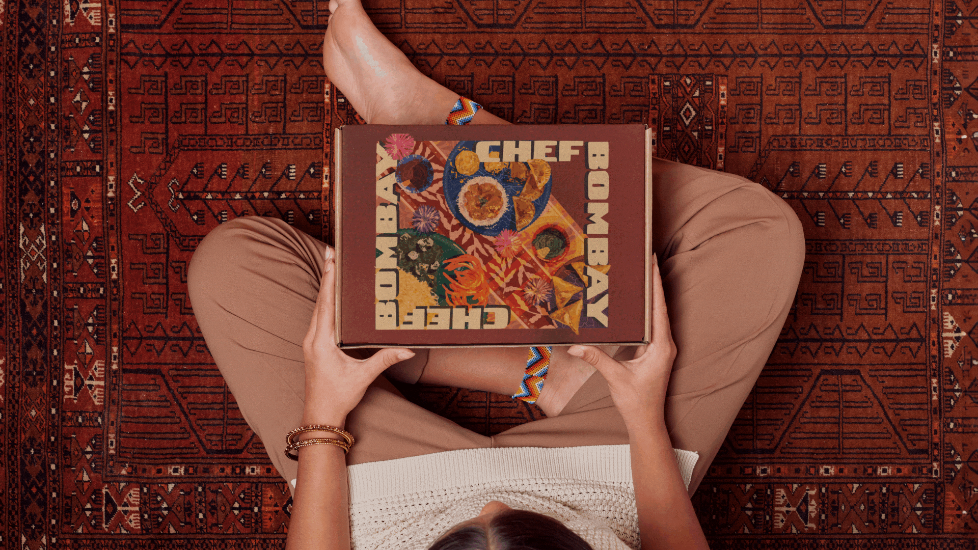 A person sitting cross-legged on a patterned rug, holding a cardboard box. The person's left arm is visible, wearing bracelets, and their right hand holds the box. They are wearing beige pants, a white top, and a multicolored bracelet.
