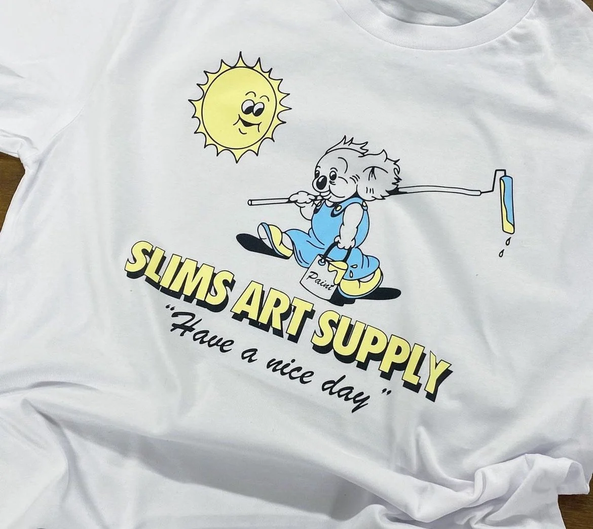 White t-shirt with cartoon koala holding a paint roller and bucket under a smiling sun, text reads "SLIMS ART SUPPLY 'Have a nice day'"