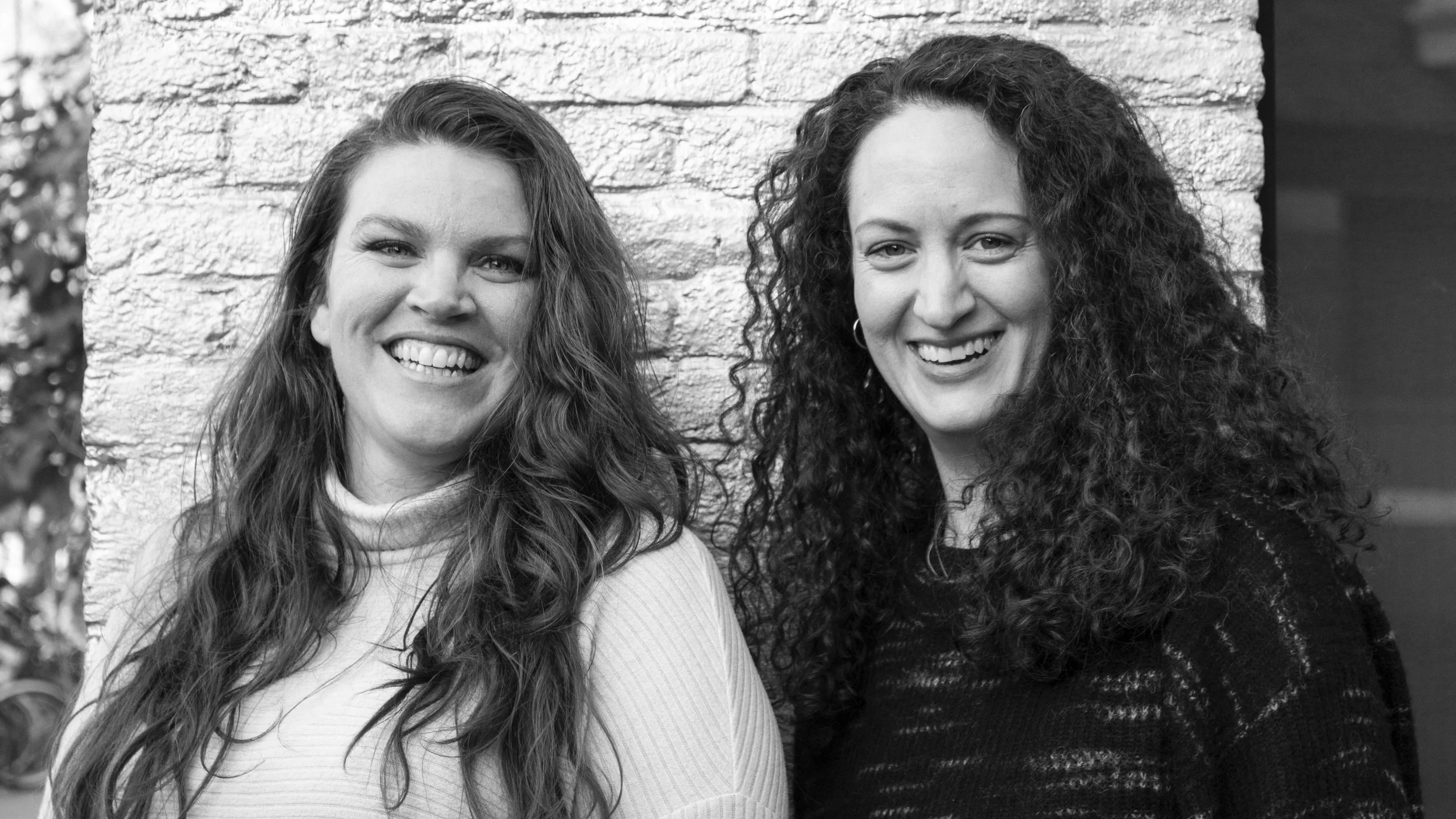 Two women smiling and standing close together outdoors against a brick wall, one with long wavy hair wearing a light turtleneck, the other with long curly hair wearing a dark sweater.