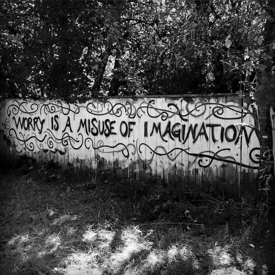 A graffiti mural on a wall reads, 'Worry is a misuse of imagination,' surrounded by decorative swirls and set against a backdrop of trees and foliage.