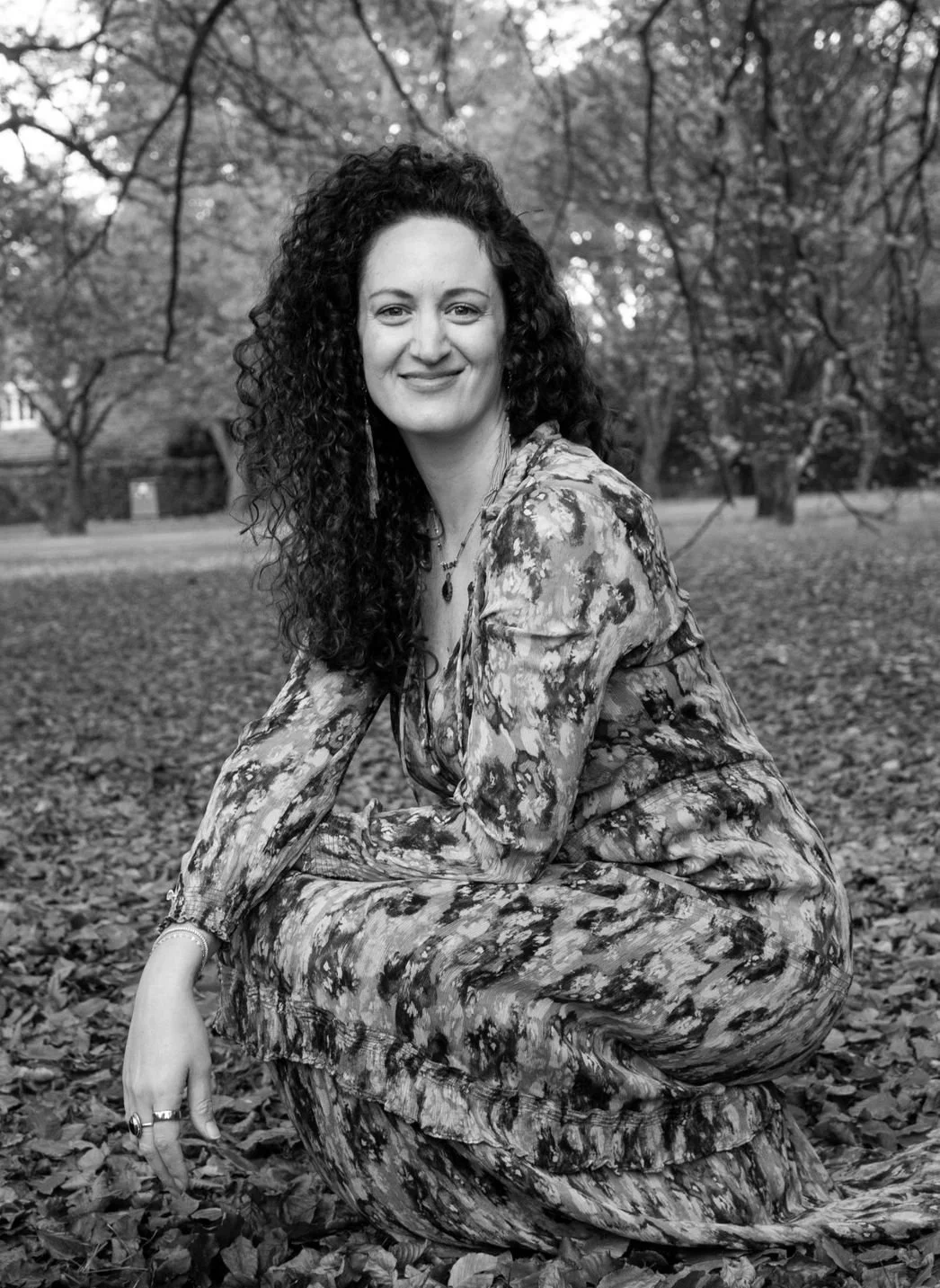 A woman with curly hair wears a floral dress and sits on the ground covered with fallen leaves in a park, smiling at the camera, surrounded by trees.