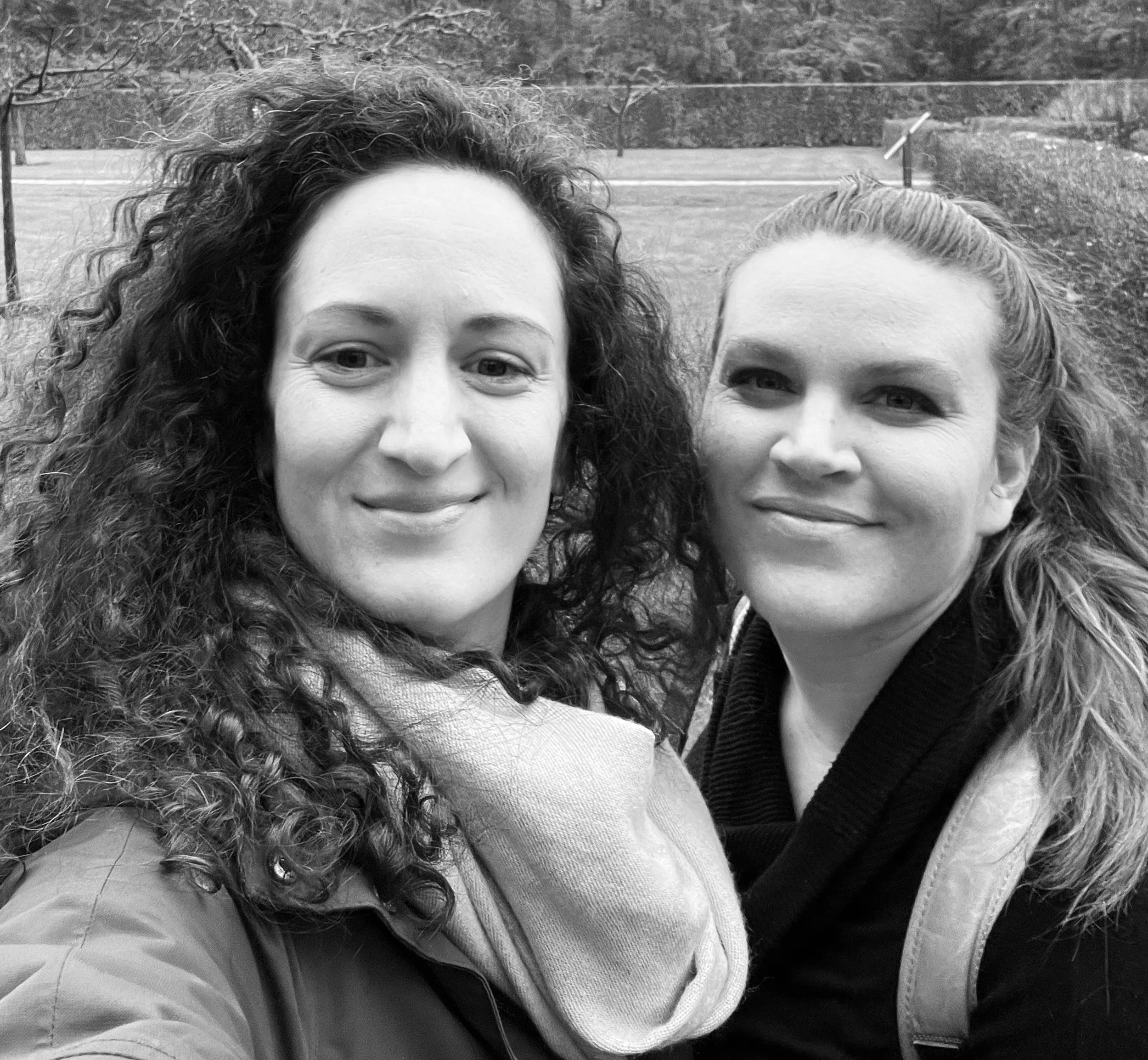 Black and white photo of two women taking a selfie outdoors, smiling, with trees and a park in the background.