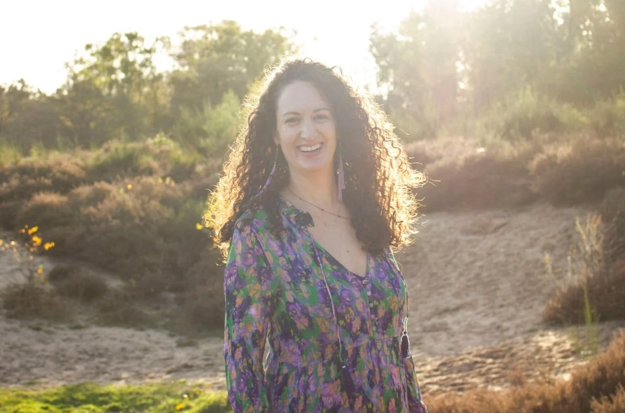 Woman with curly dark hair smiling outdoors in natural sunlight with trees and bushes in the background, wearing a colorful patterned dress.