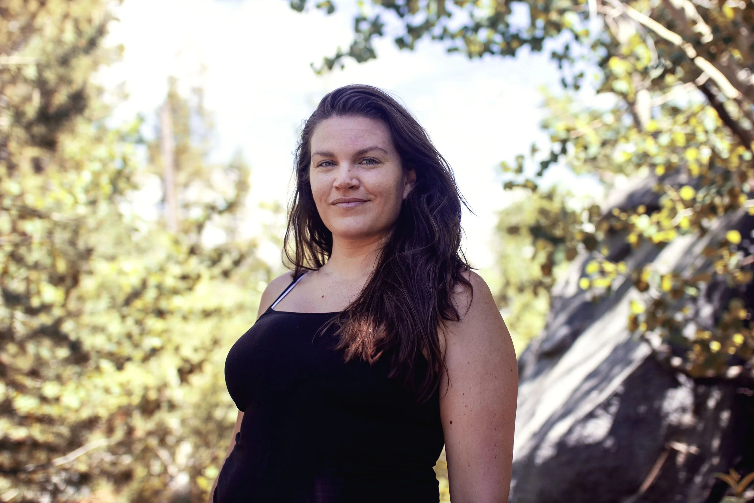A woman with long dark hair standing outdoors on a sunny day, wearing a black tank top, with trees and rocks in the background.
