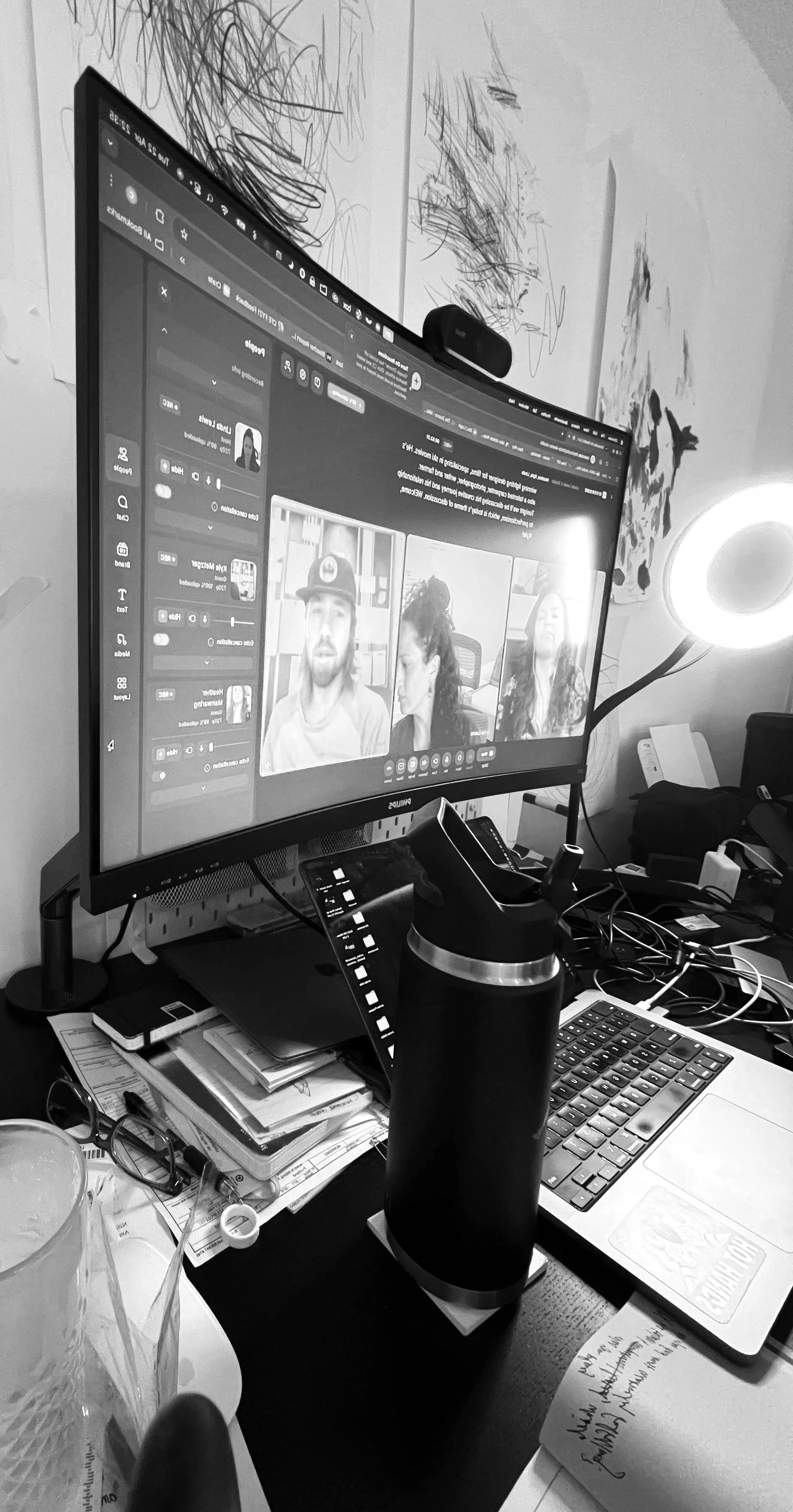 A computer monitor displaying a video conference with three participants, surrounded by various office supplies, a laptop, a water bottle, and a lamp on a cluttered desk.