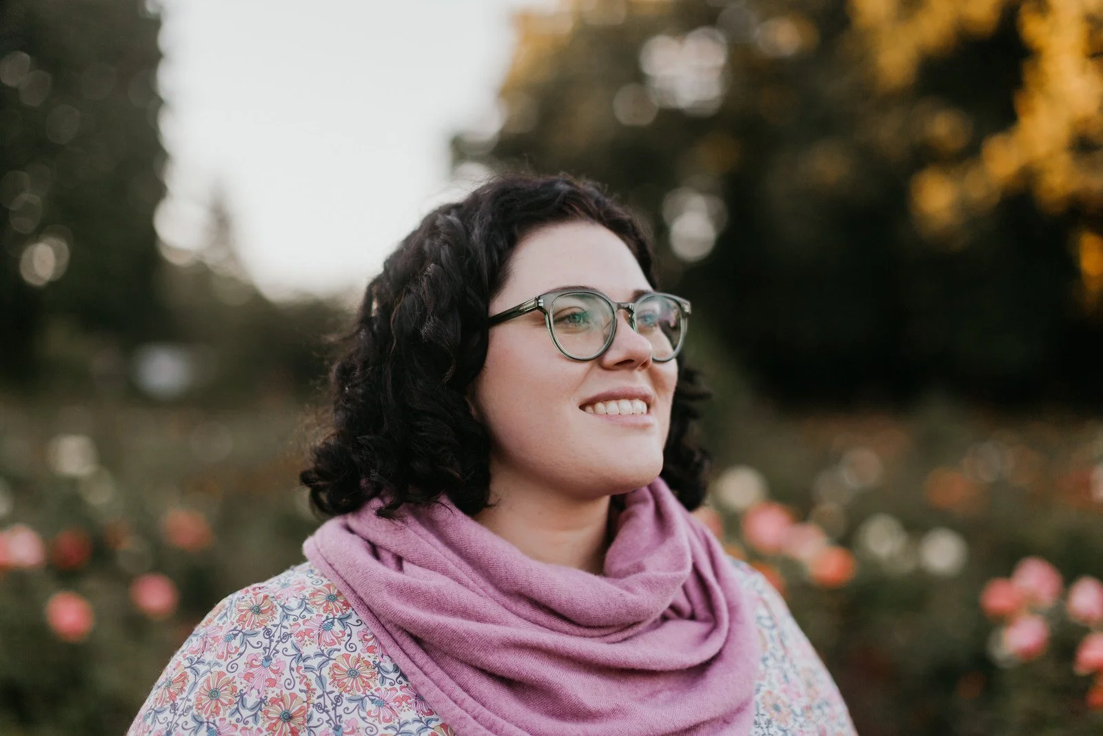 A woman with dark curly hair and glasses, wearing a pink scarf and a floral top, smiling outdoors during sunset with blurred pink flowers and trees in the background.