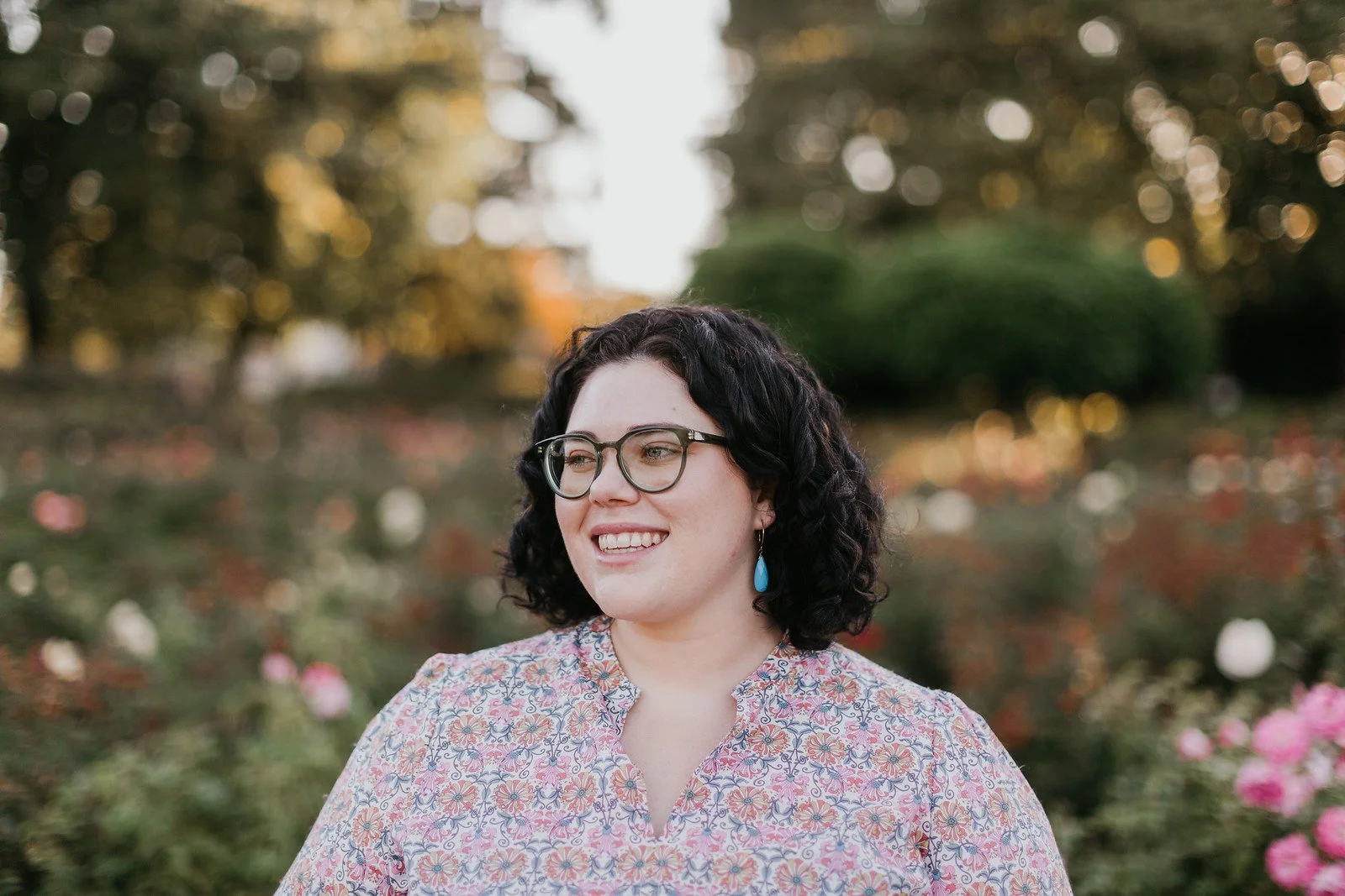 A young woman with dark curly hair, glasses, and earrings, smiling outdoors in a park or garden during autumn, with trees and pink flowers in the background.