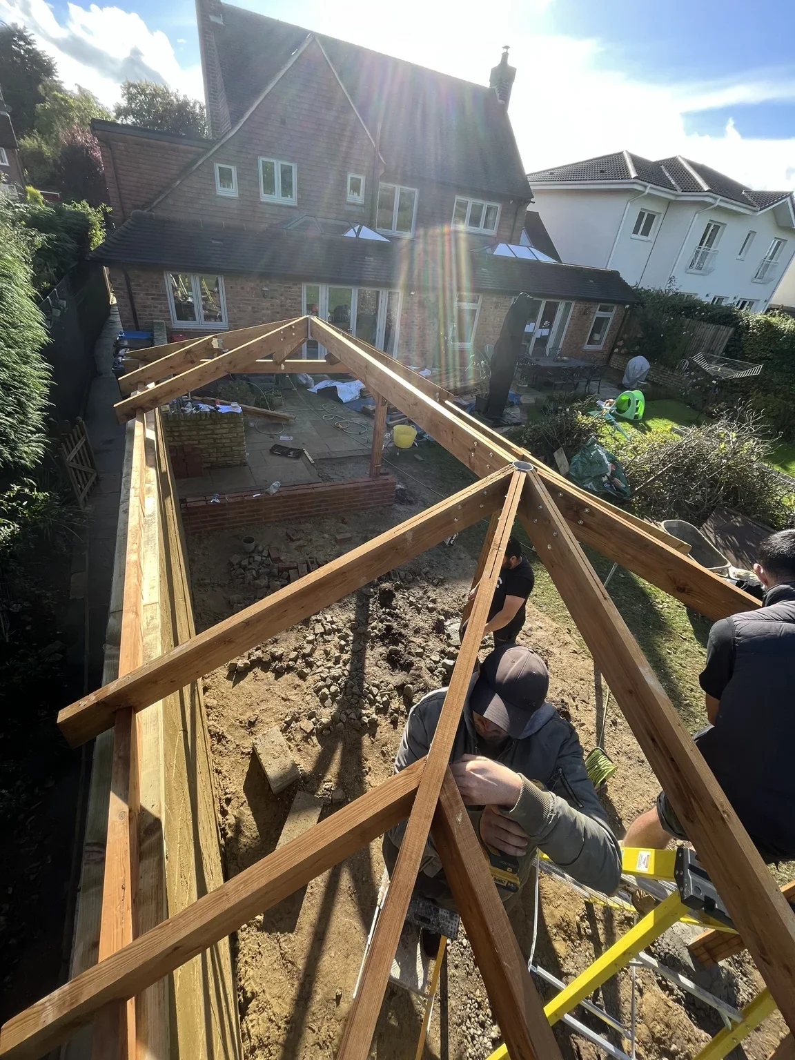 Timber shelter in progress — the roof framework is being assembled above the brick base before the covered seating area is completed.