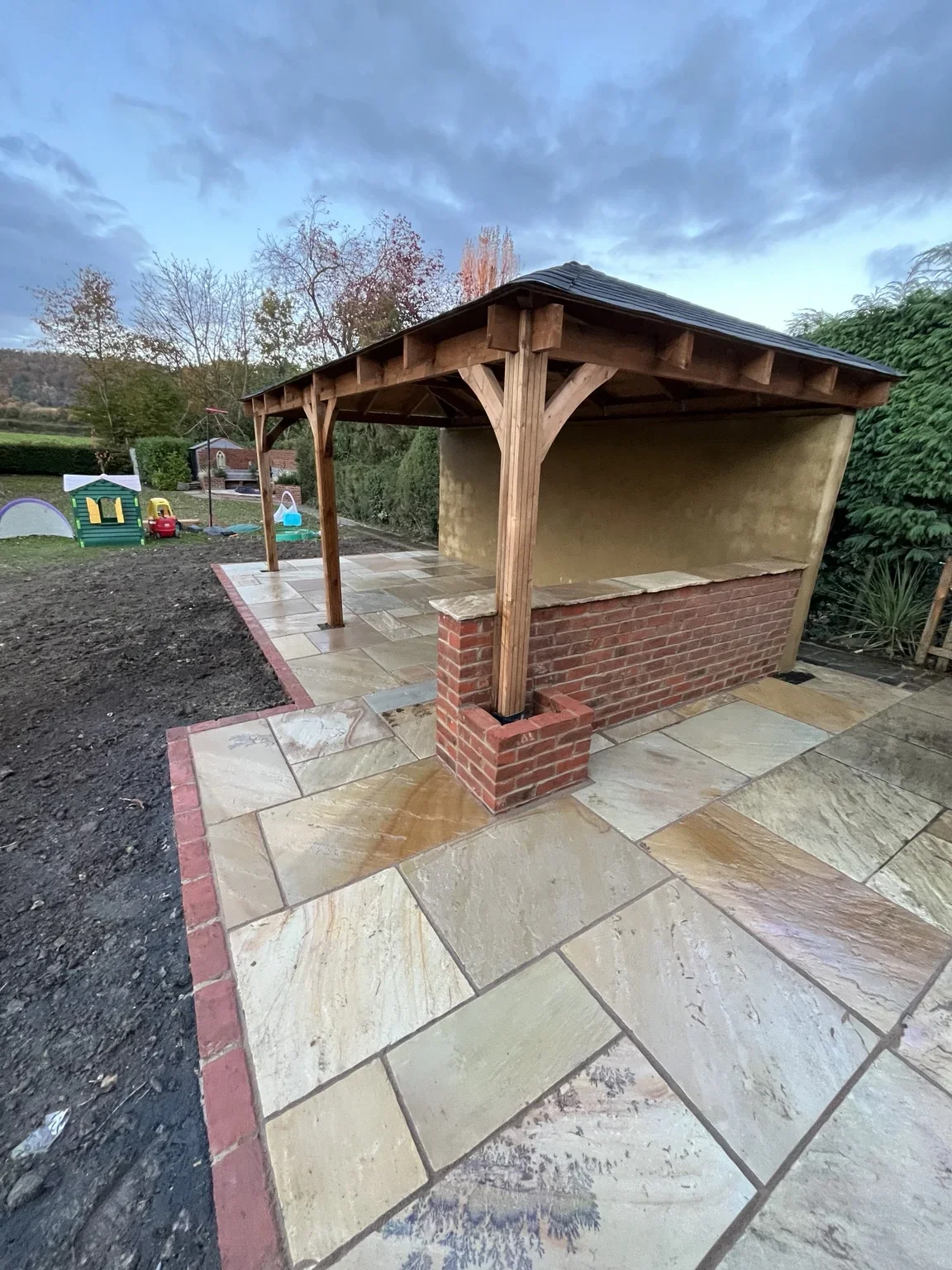 Covered garden seating area — a timber roof and posts sit above a sandstone floor, with low brick walls and matching edging around the structure.