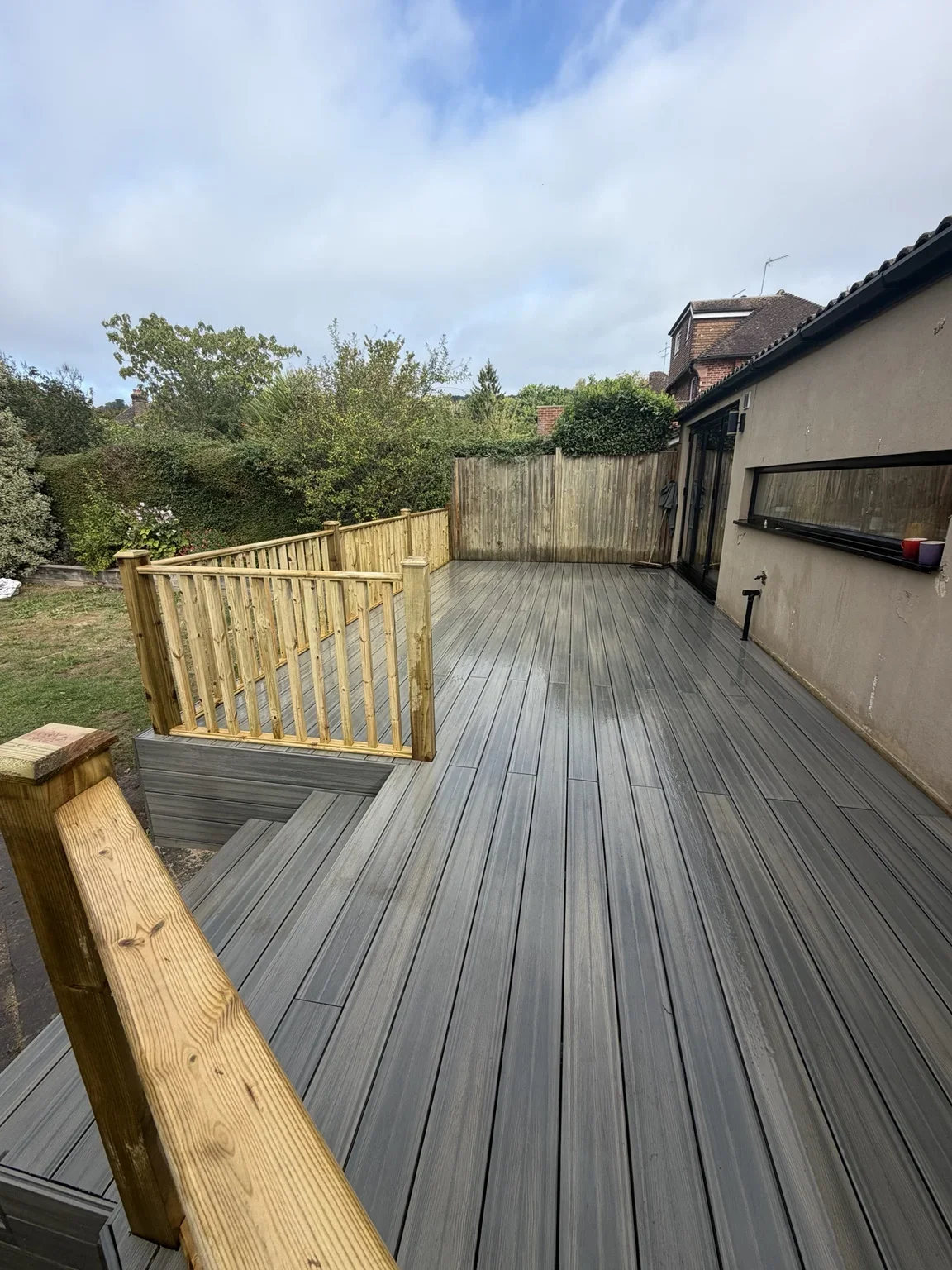 Composite decking detail — parallel grey boards run beside the house, while timber balustrades enclose the raised deck and its steps.