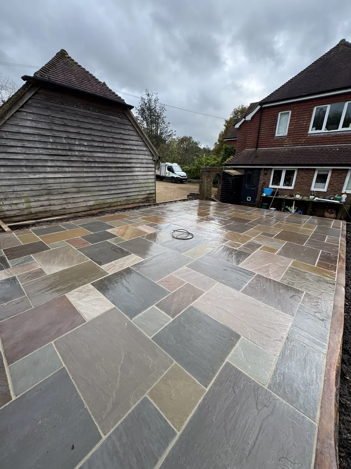 Sandstone patio — a mix of natural stone colours laid in a varied-size pattern, bordered by brick edging beside the timber garden building.