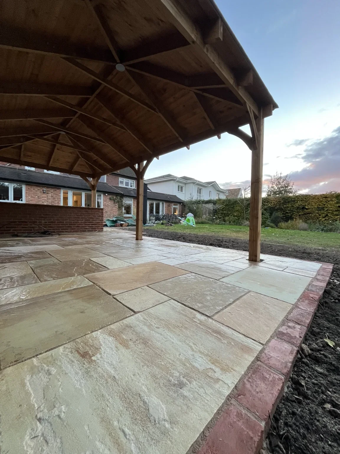 Inside the covered seating area — sandstone paving continues beneath the timber roof, with brick edging marking the open side of the shelter.