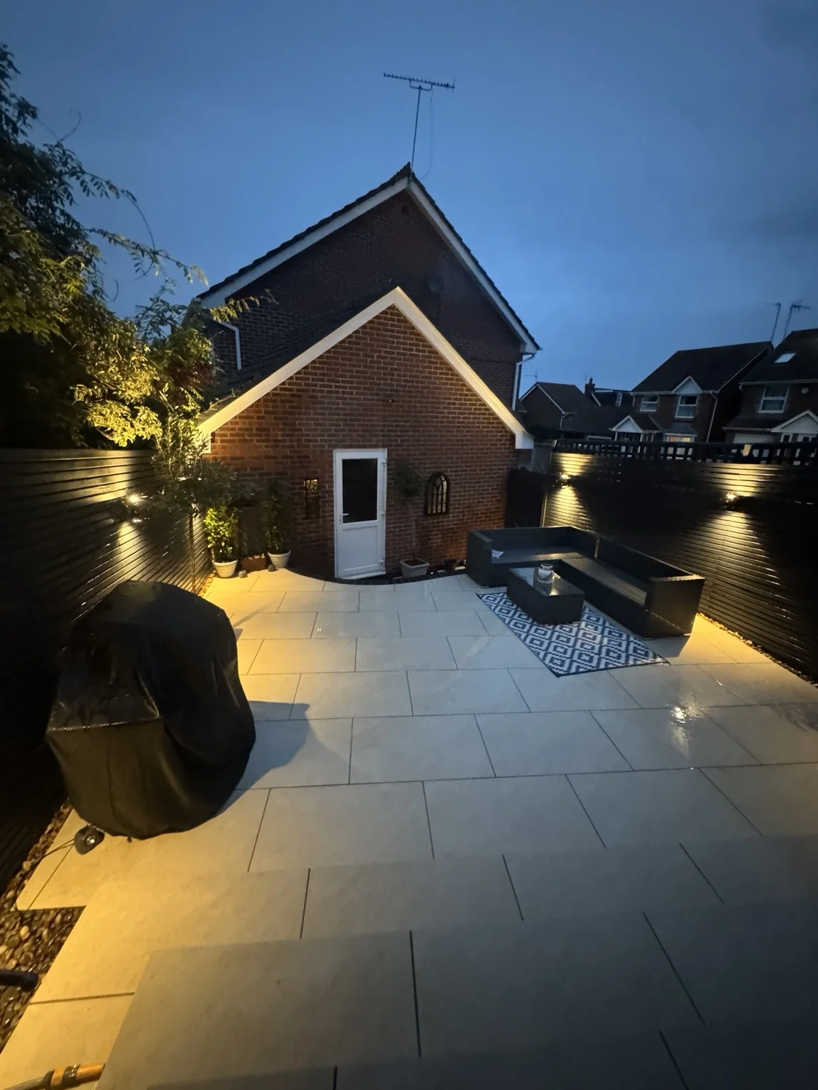 Patio at dusk — warm lighting picks out the paving and steps around the garden building, showing how the space looks in the evening.