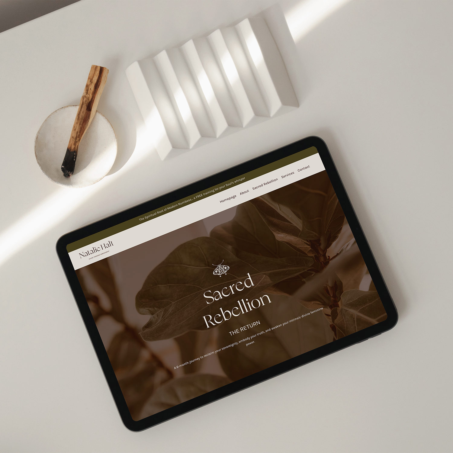 Une tablette partiellement visible affichant une page web intitulée « Sacred Rebellion » sur fond de feuilles, posée sur une surface blanche. À proximité se trouve un petit plat en céramique sur lequel repose une cuillère en bois, ainsi qu'un élément décoratif blanc.