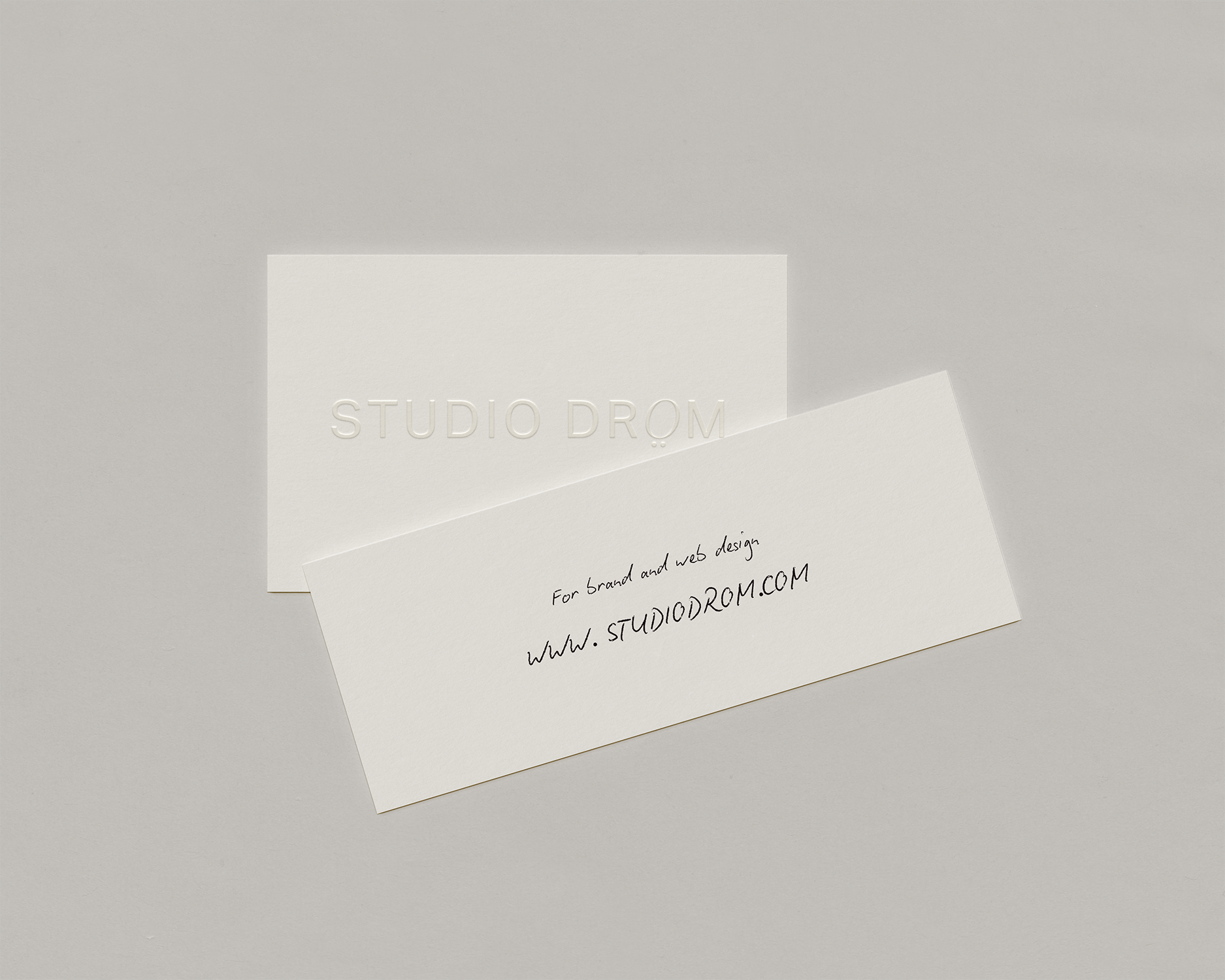 Une carte de visite blanche avec l'inscription « STUDIO DROM » en relief et une note manuscrite indiquant l'adresse du site web www.studiodrom.com sur un fond gris clair.