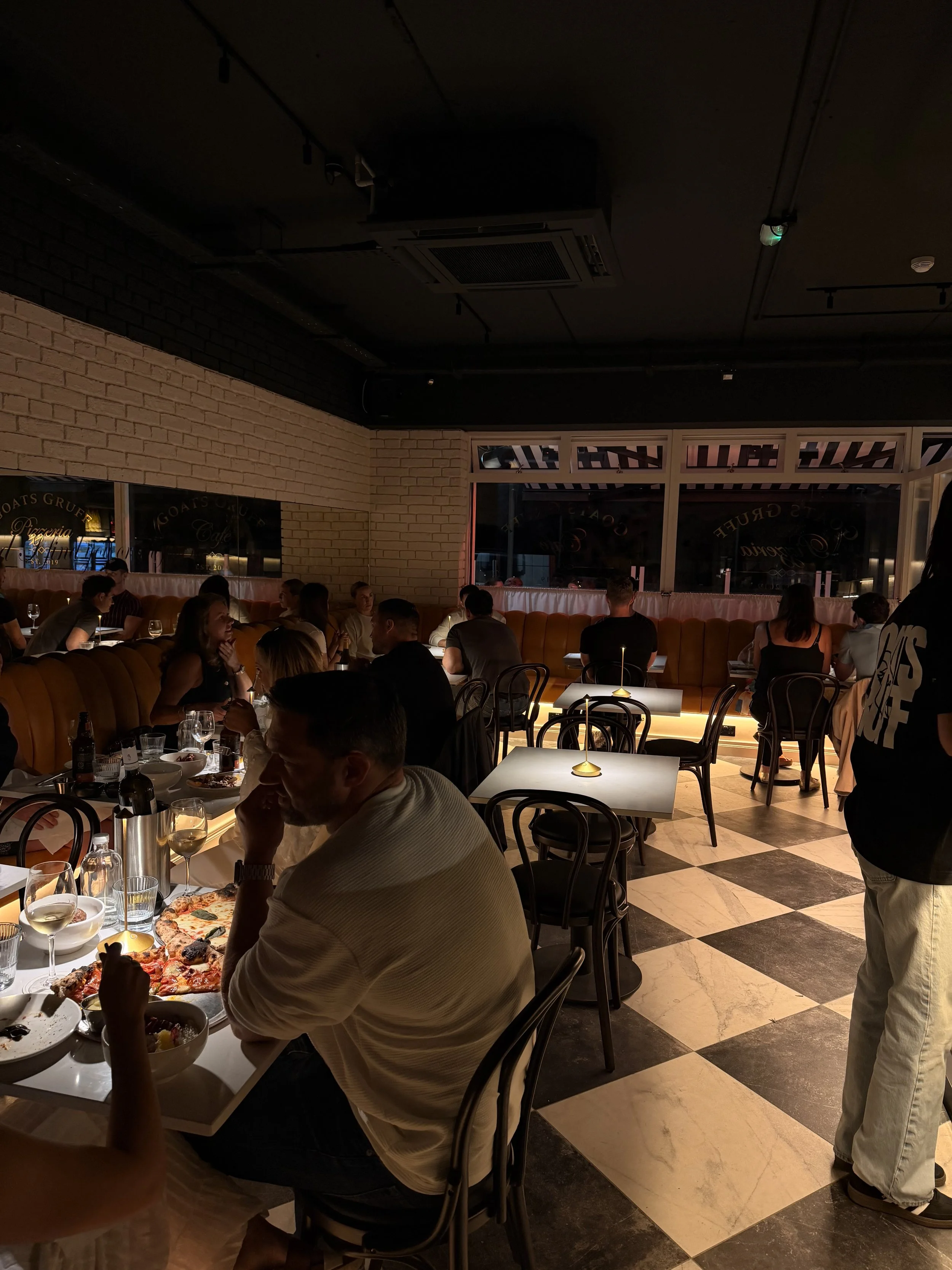 People dining in a warmly lit restaurant with brick walls, glass windows, and checkered marble floor tiles, enjoying pizza and drinks.