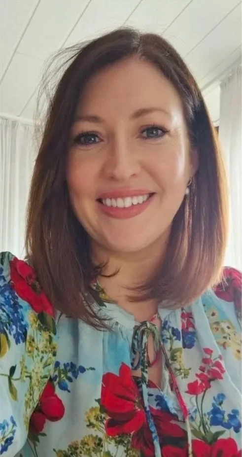 A woman with shoulder-length brown hair and blue eyes, smiling, wearing a floral blouse with red, yellow, blue, and green flowers.