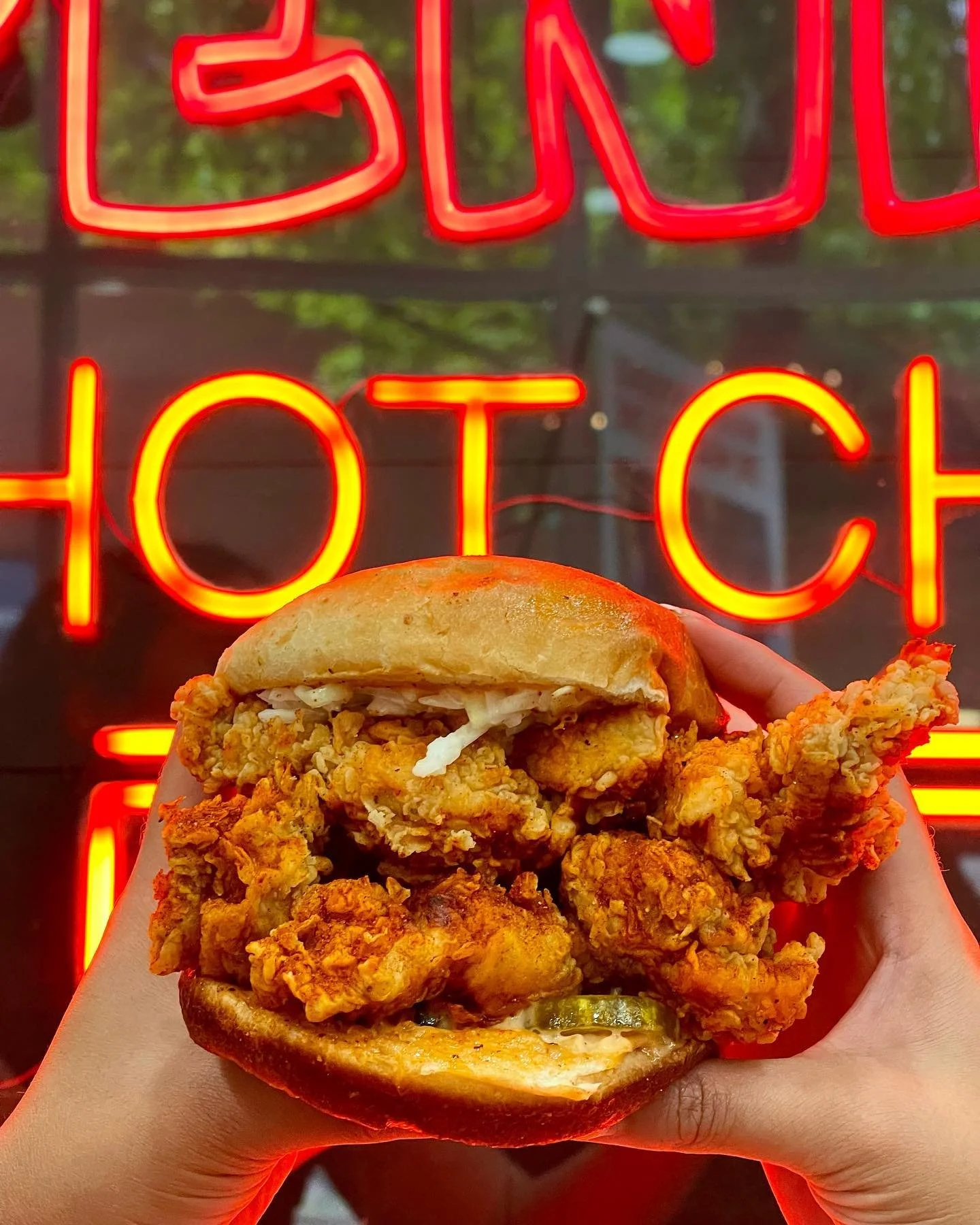 Close-up of a fried chicken sandwich with pickles, lettuce, tomato, and bun, held in front of a neon sign that says 'Eat Hot'