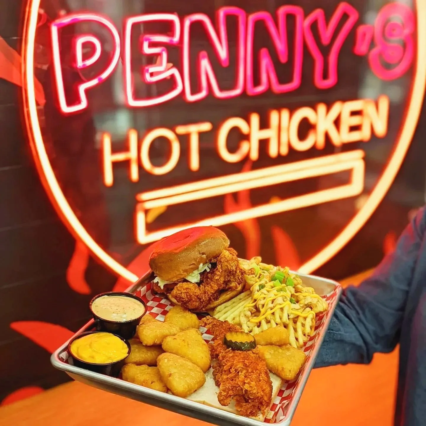 Tray of fried chicken, chicken sandwich, potato nuggets, fried fish, corn, and dipping sauces in front of neon sign that says "Penny's" and "Hot Chicken".