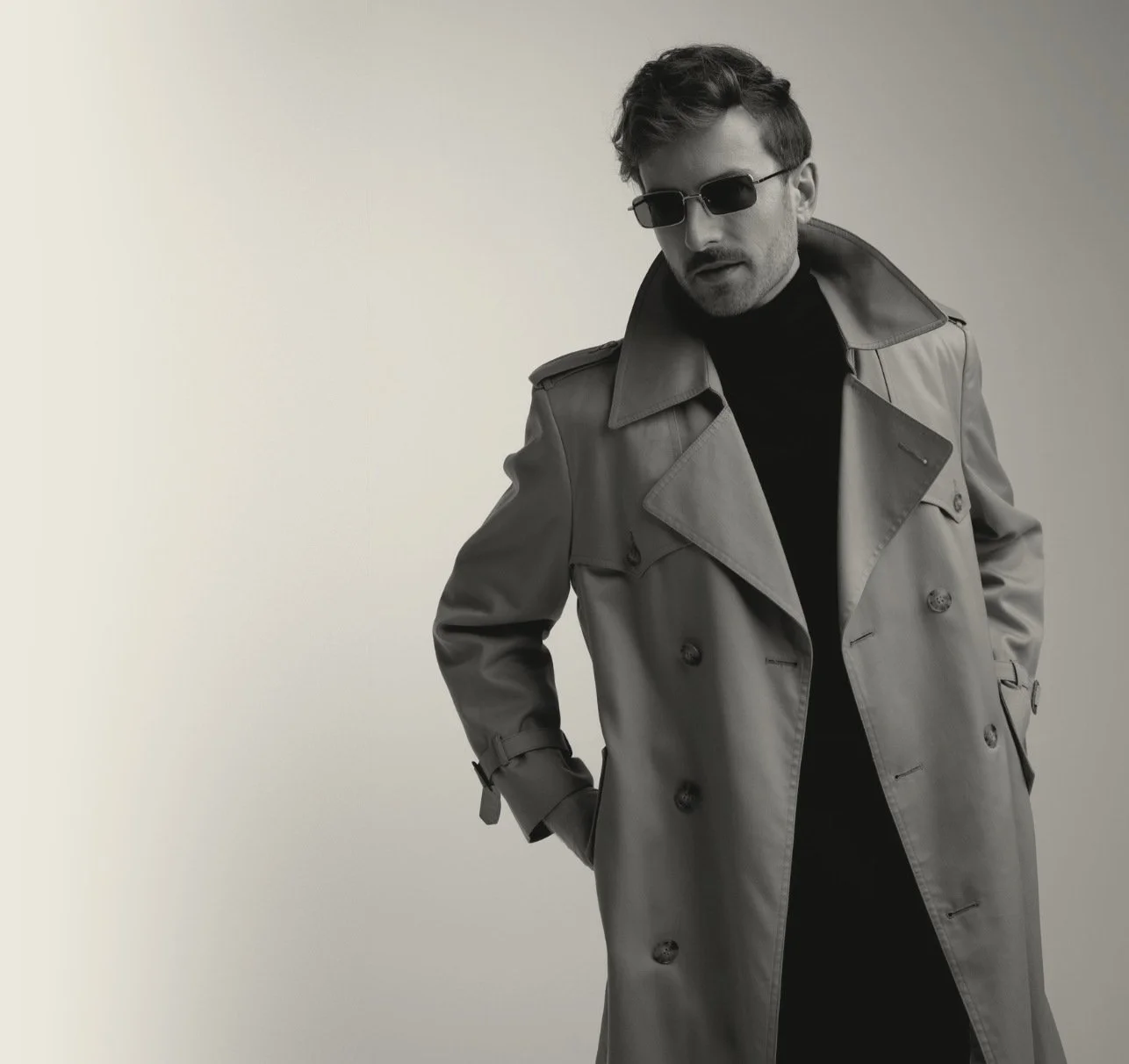 Black-and-white photo of Evan Nathaniel Grim wearing sunglasses, a trench coat, and a dark turtleneck, posing with his hands in pockets against a plain light background.