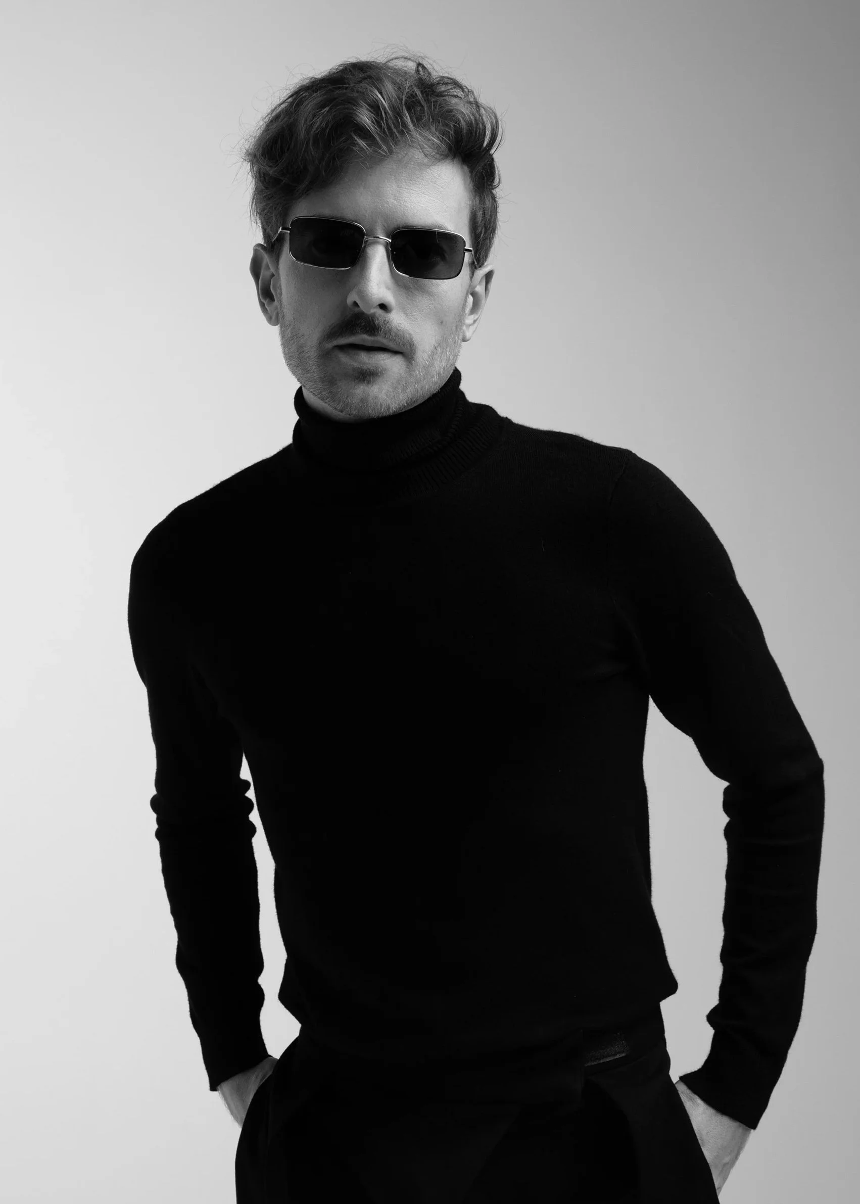 A black-and-white photo of Evan Nathaniel Grim wearing a black turtleneck, sunglasses, and with tousled hair, standing with his hands in his pockets against a plain background.