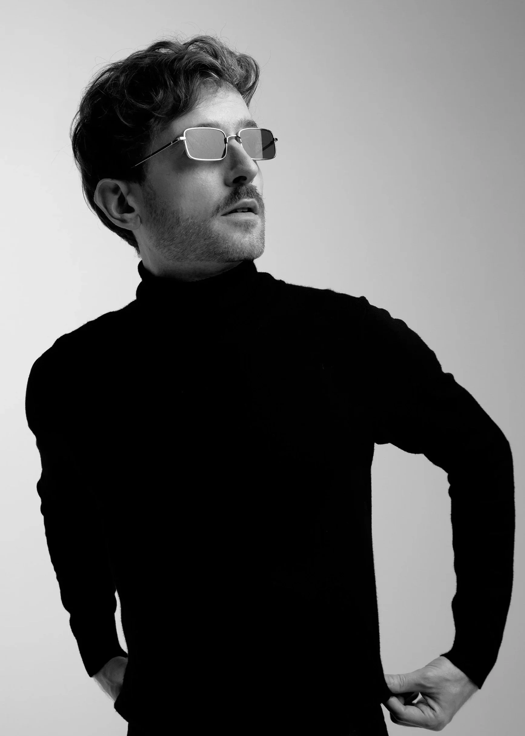 Black and white photo of Evan Nathaniel Grim with wavy hair wearing sunglasses and a black turtleneck, standing against a plain background.