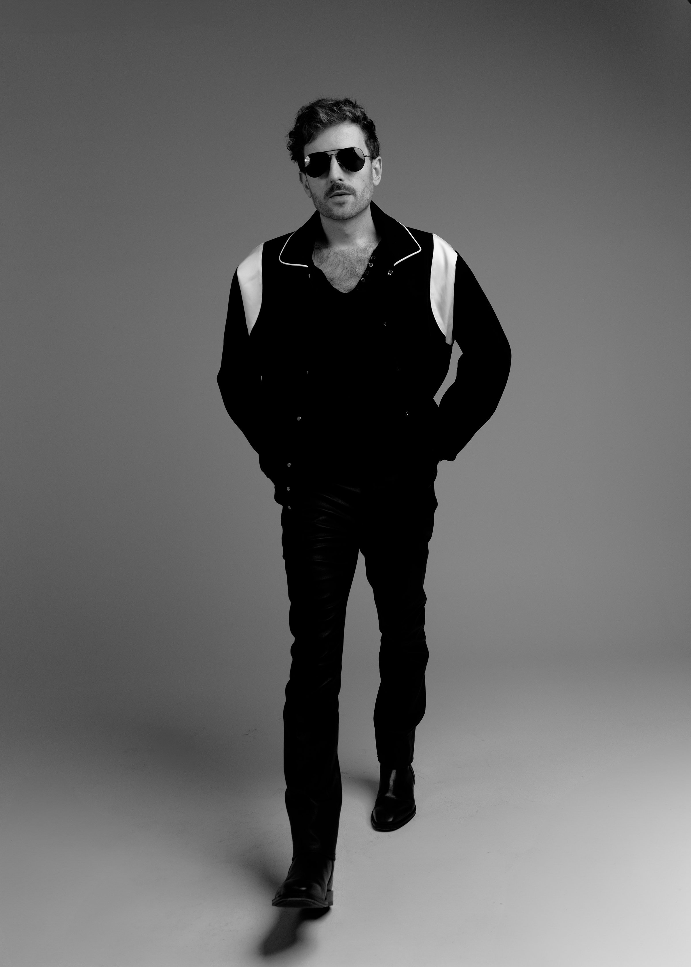 Evan Nathaniel Grim walking towards the camera wearing sunglasses, a black shirt, and black pants, in a black-and-white studio setting.