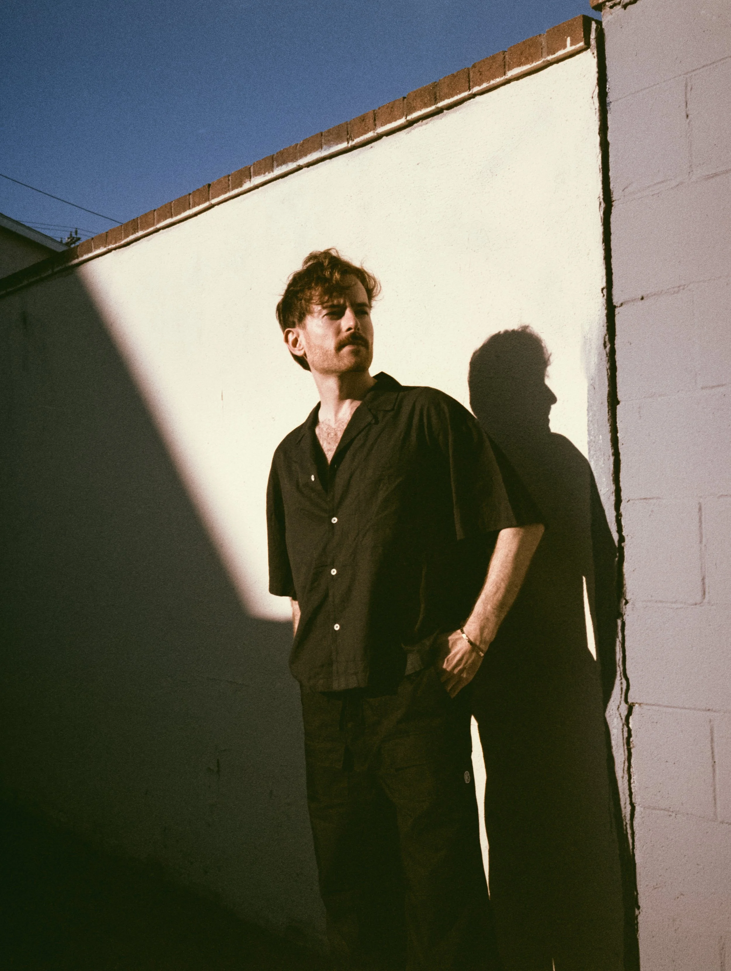 Evan Nathaniel Grim wearing a black shirt stands outdoors near a white wall, with his shadow cast on the wall, during twilight or evening.