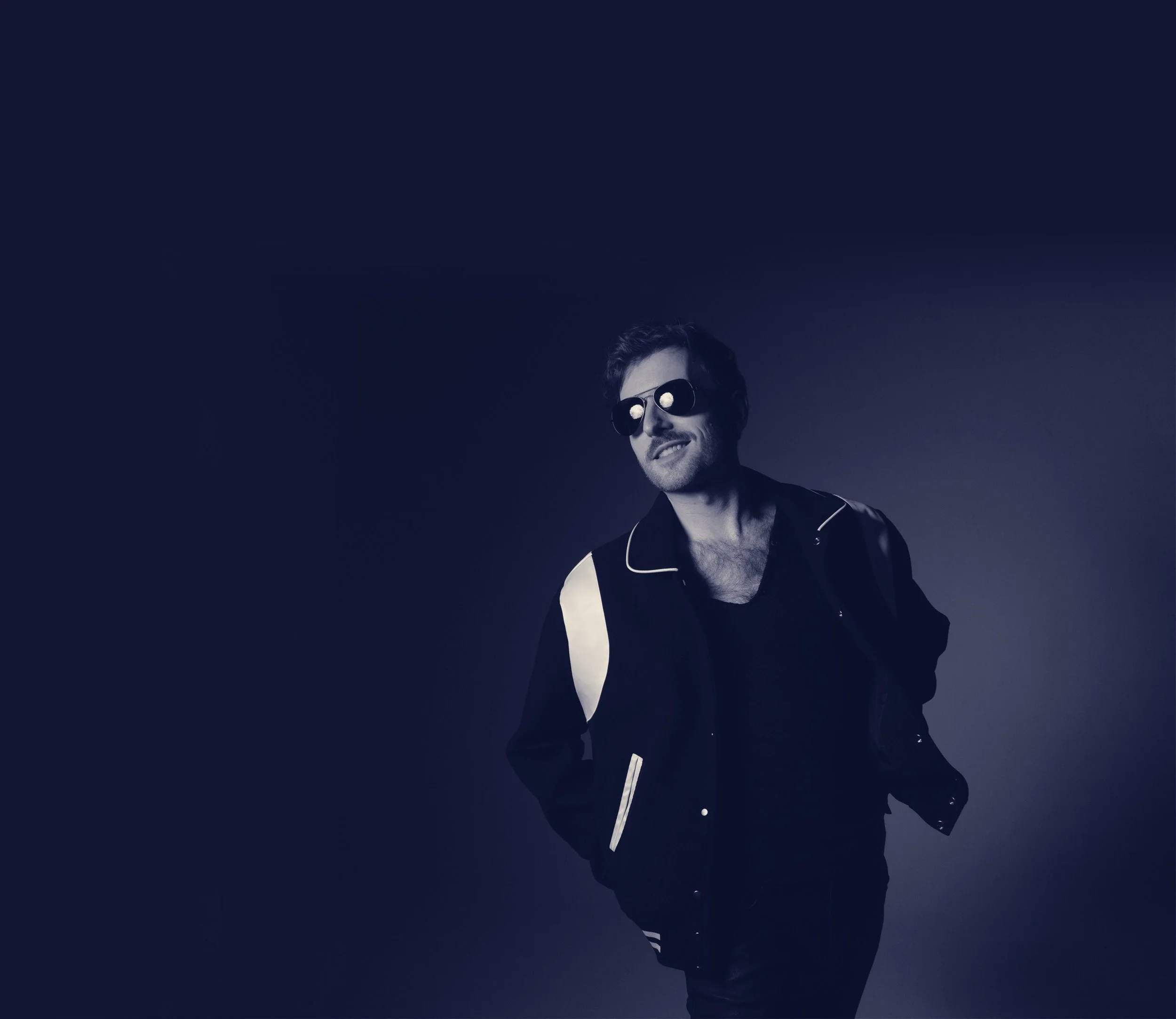 Evan Nathaniel Grim smiling wearing sunglasses and a black jacket with white accents, standing against a dark background.