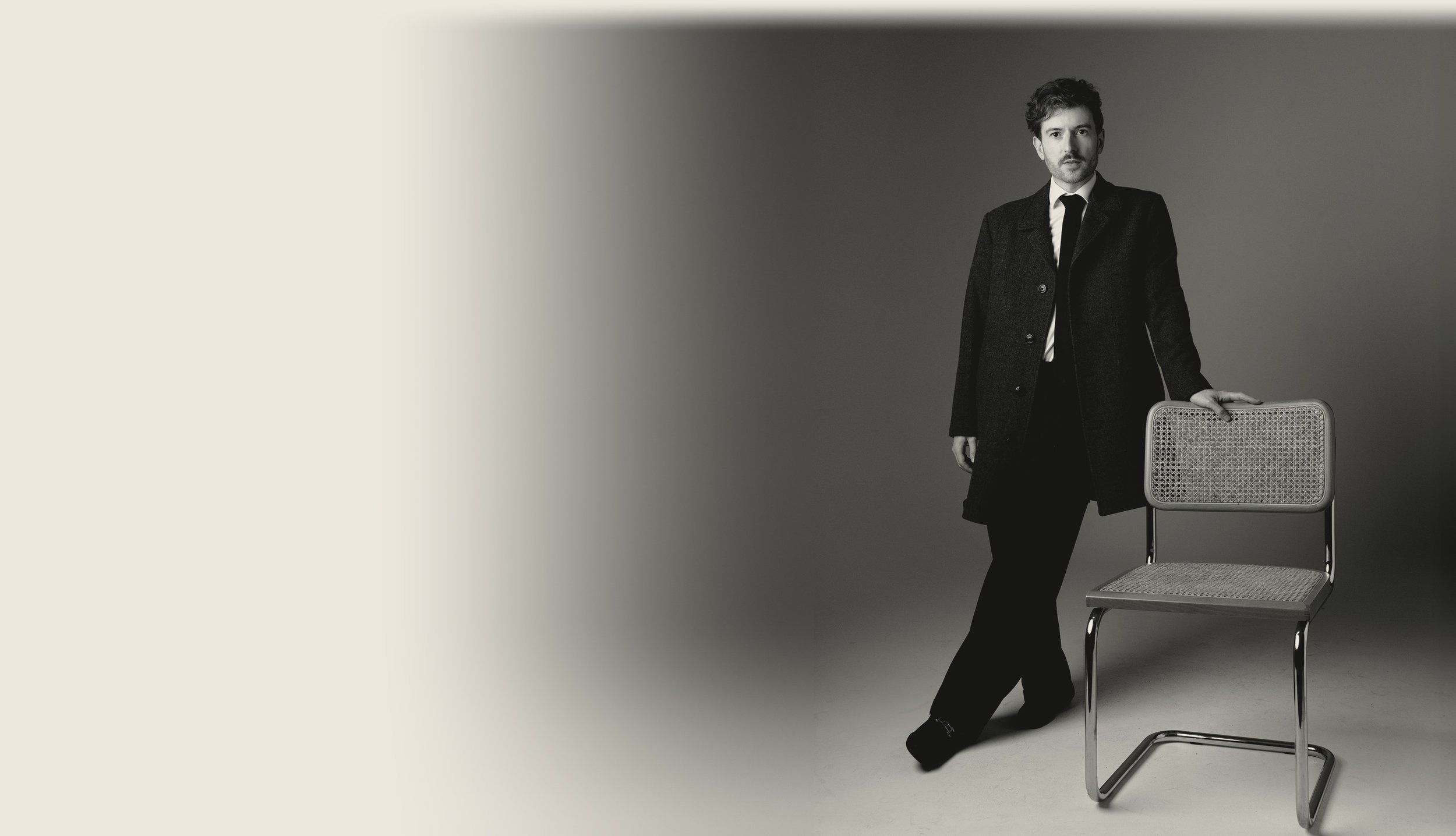 Evan Nathaniel Grim wearing a dark coat and tie standing next to a chair with a cane-like frame and woven seat and backrest in a studio setting.