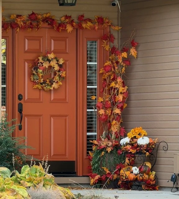 An autumn-themed front porch with a coral-colored door, a wreath made of colorful fall leaves, and a decorative display of pumpkins and gourds surrounded by fall foliage.