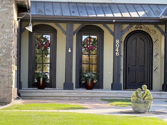 Front porch of a house with two large windows with flower boxes, a black arched door, a decorative stone sculpture of two children sitting in a nest, and the street address 8046.
