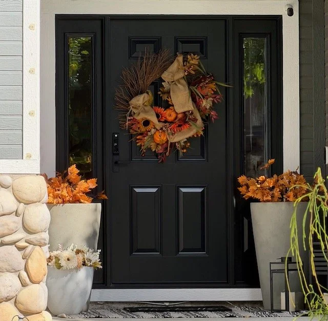 Front door decorated with an autumn-themed wreath and two large planters filled with orange and yellow fall leaves.