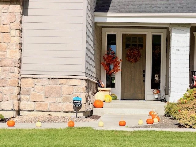 Front porch of a house decorated for fall, with pumpkins and gourds on the steps and yard, autumn wreath on the door, and small trees with fall foliage.