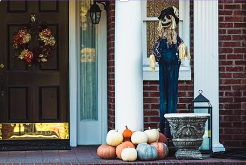 Front porch with a Halloween decoration of a skeleton wearing a blue denim outfit and a black hat, standing next to a pile of pumpkins of various colors, a decorative stone planter, and a lantern, in front of a brick house.