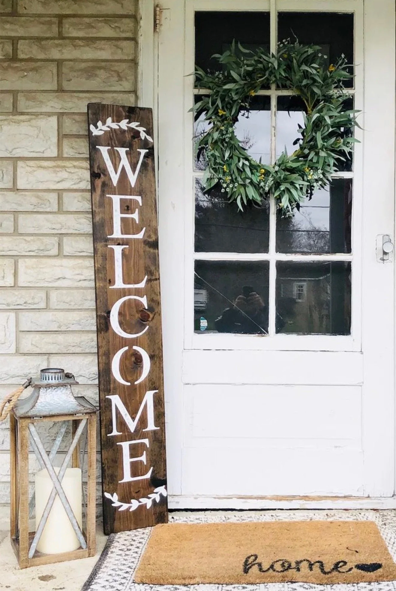 A decorative front door area with a green wreath on a white door, a tall wooden welcome sign leaning against the wall, a lantern with a candle inside, and a doormat that says 'home'.