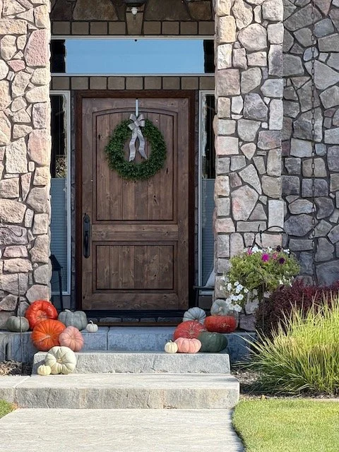 Front door decorated with a wreath, surrounded by pumpkins and gourds, with a potted plant on the right side and a stone facade.