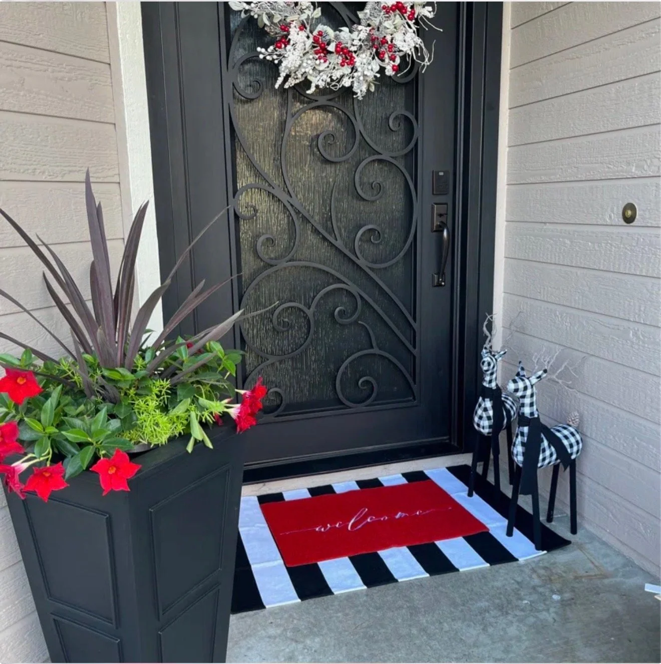 Front door decorated with a red and white holiday wreath, a striped black-and-white rug with a red welcome mat, two black-and-white checkered reindeer figurines, and a tall planter with red flowers and green plants beside the door.