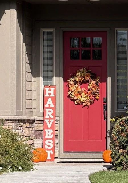 Red front door decorated with a fall wreath, surrounded by pumpkins and a harvest sign.