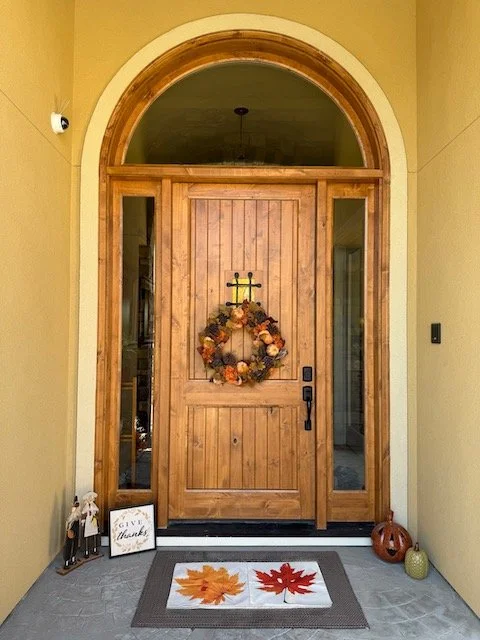 Front door with a wooden door and an arched transom window above it, decorated with a fall-themed wreath made of flowers, leaves, and pumpkins. Fall-themed decorations include a small pumpkin and a gnome figurine on the porch, along with a sign that
