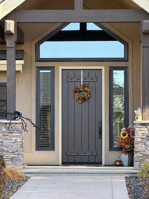Front door with a wreath, flanked by two tall windows with blinds, and topped with a large triangular and rectangular window. There are stone columns on either side of the steps leading to the door, with sunflower and autumn-themed decorations.