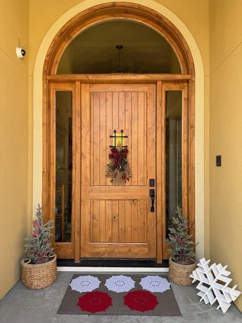 Front door with decorative wreath, potted Christmas trees on each side, and holiday decorations on a welcome mat.