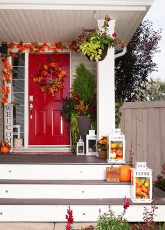 Front porch decorated for fall with pumpkins, lanterns, and autumn leaves, featuring a bright red door and potted plants.
