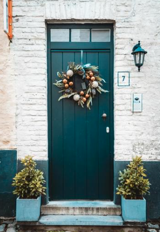 Blue front door with a holiday wreath, flanked by two potted plants, on a white brick wall with house number 7 and a mailbox.